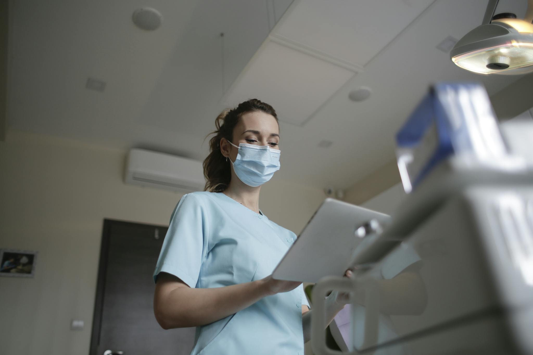 Female healthcare worker wearing a mask and holding a clipboard in a clinical environment.